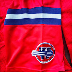 Close-up of a red athletic jersey with a blue and white chest stripe and a circular embroidered washington sentinels logo featuring stylized letters on the lower front.